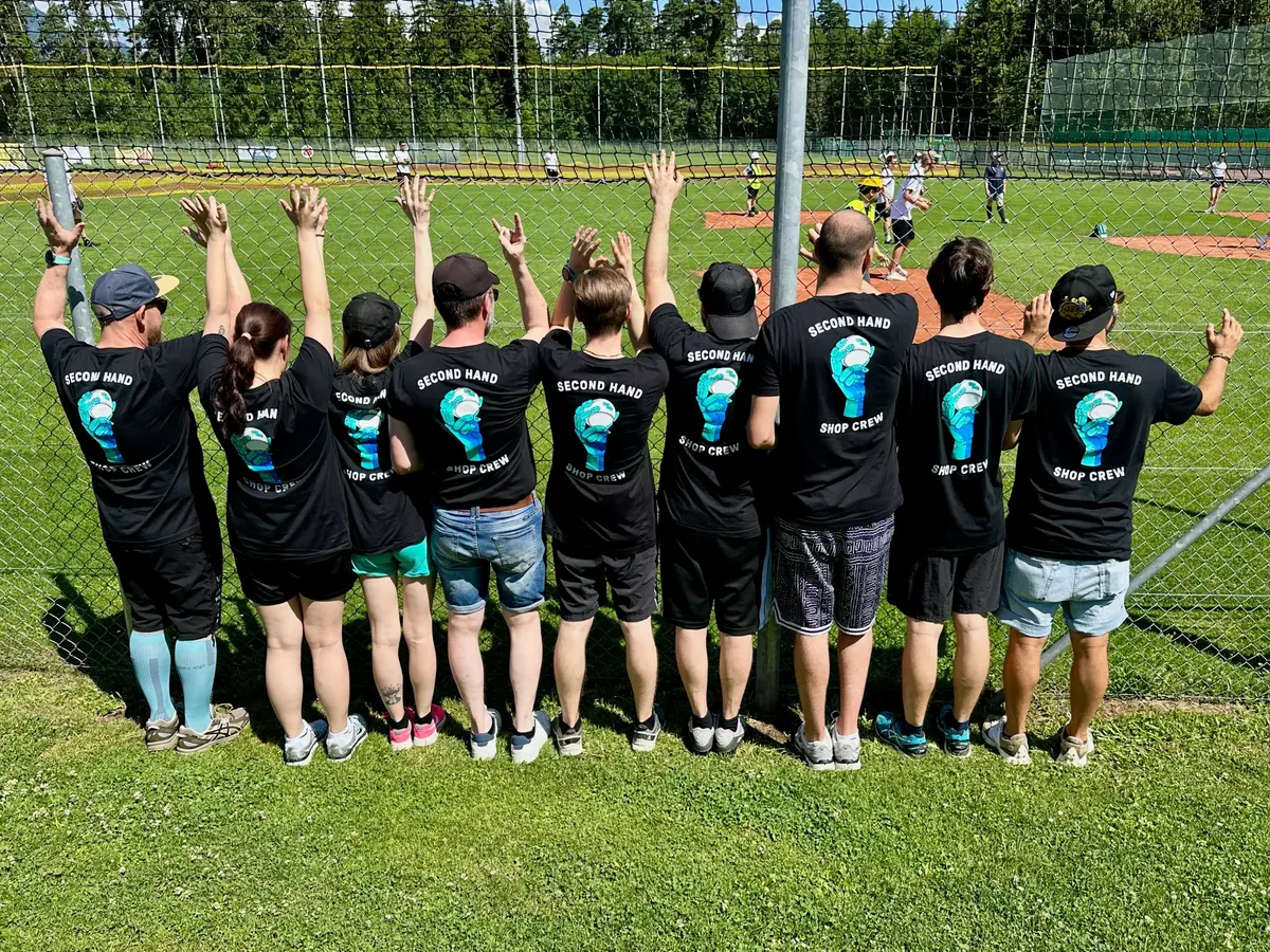Ten people in black "SECOND HAND SHOP CREW" t-shirts, seen from behind with arms raised, at a baseball field.