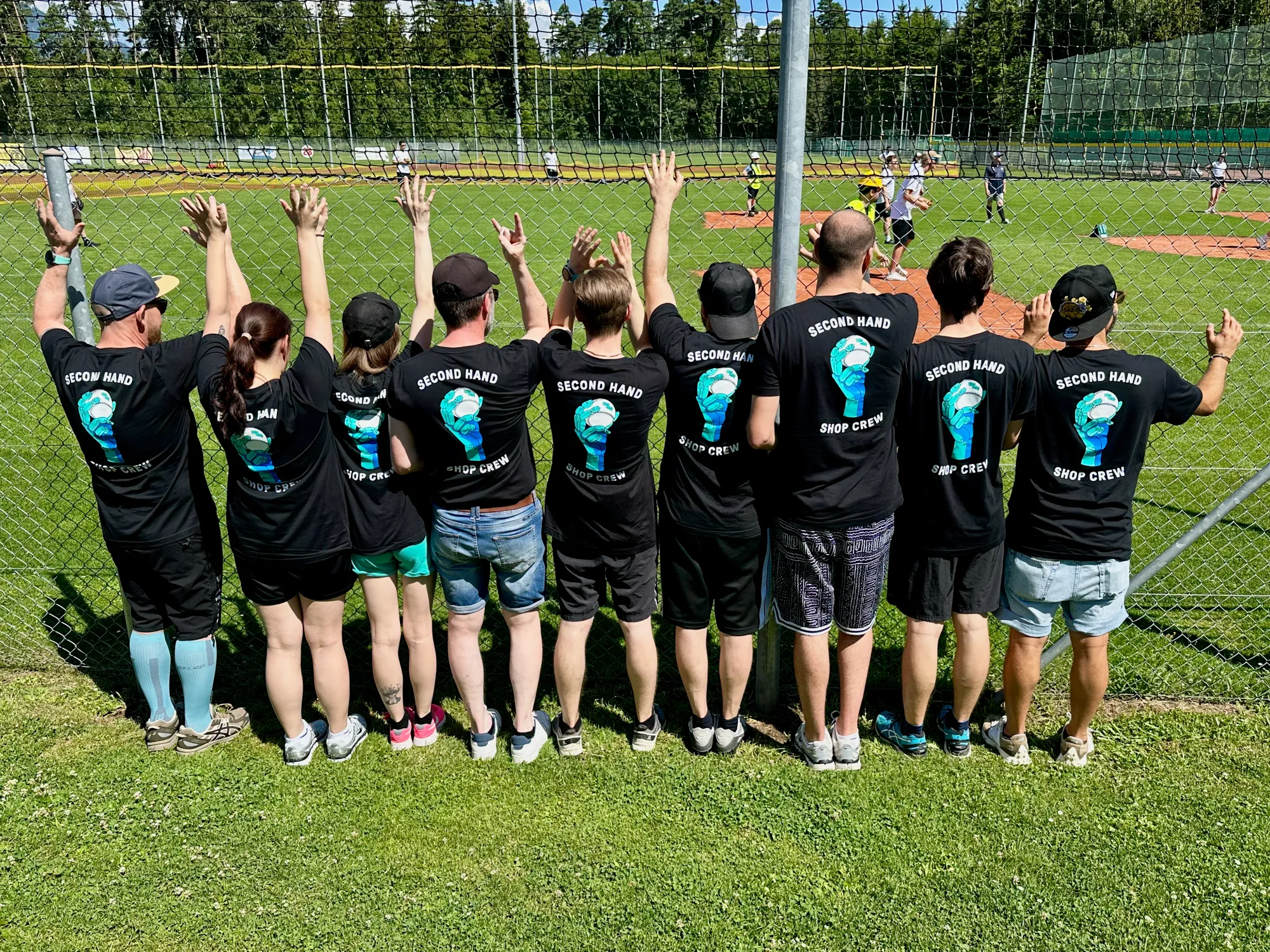 Ten people in black "SECOND HAND SHOP CREW" t-shirts, seen from behind with arms raised, at a baseball field.